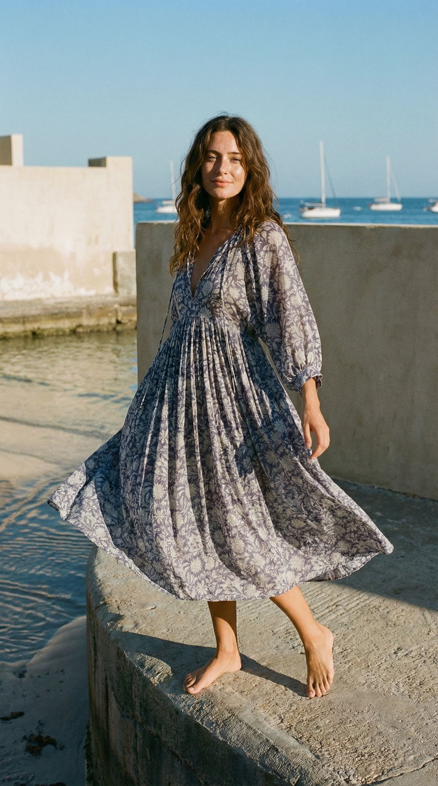 Woman in a patterned dark blue doi midi dress standing by a waterfront with boats in the background