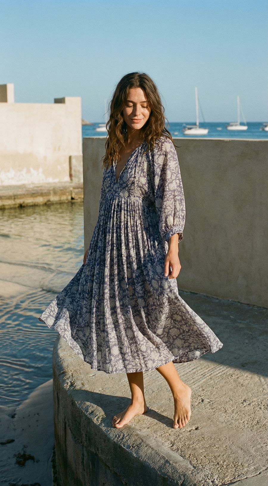 Woman in a blue patterned dress standing by a waterfront with boats in the background