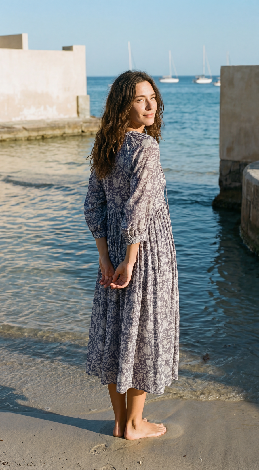Woman in a long, patterned dark blue dress standing on a beach with water and boats in the background.