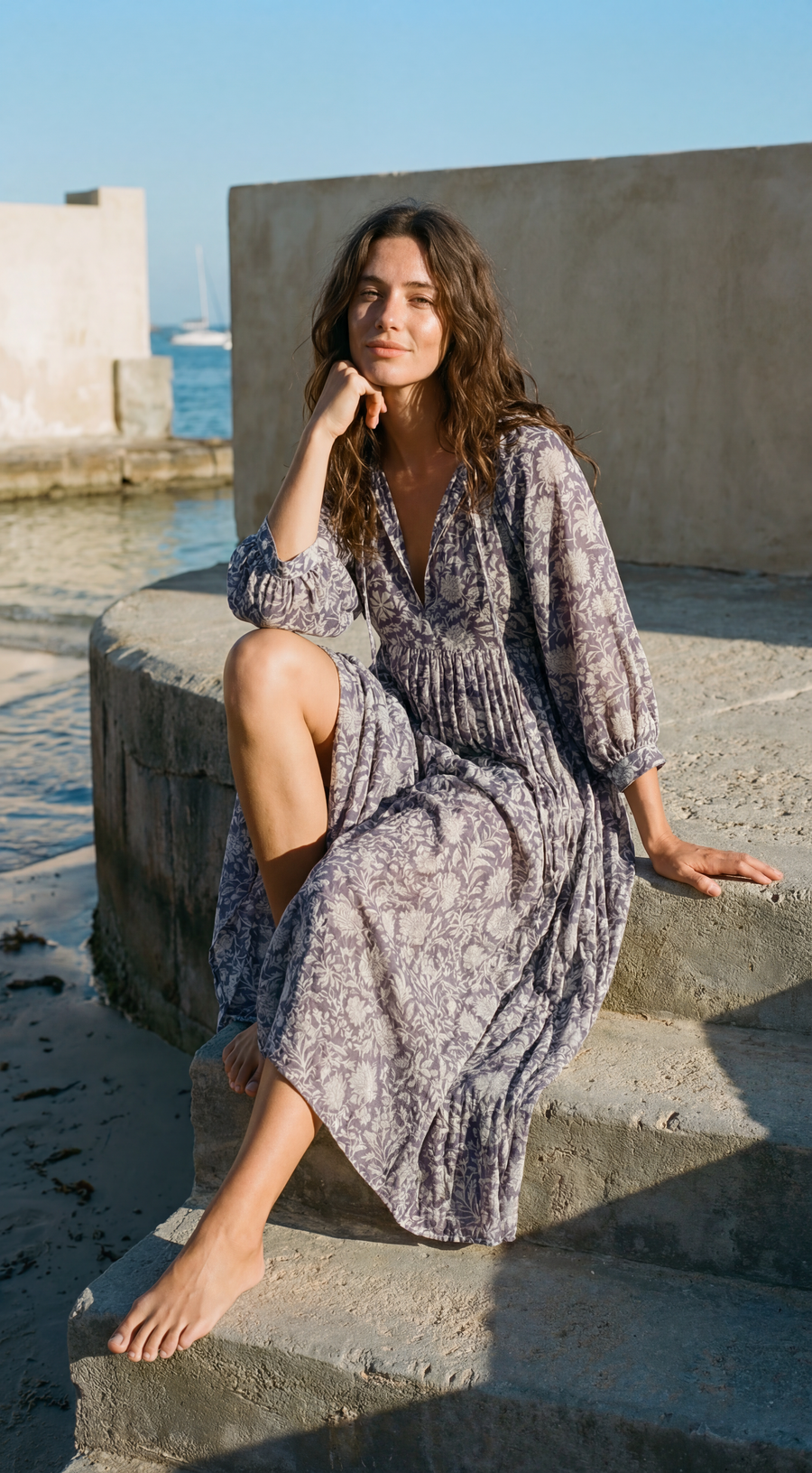 Woman in a patterned ssea dark blue dress sitting on concrete steps by a waterfront.